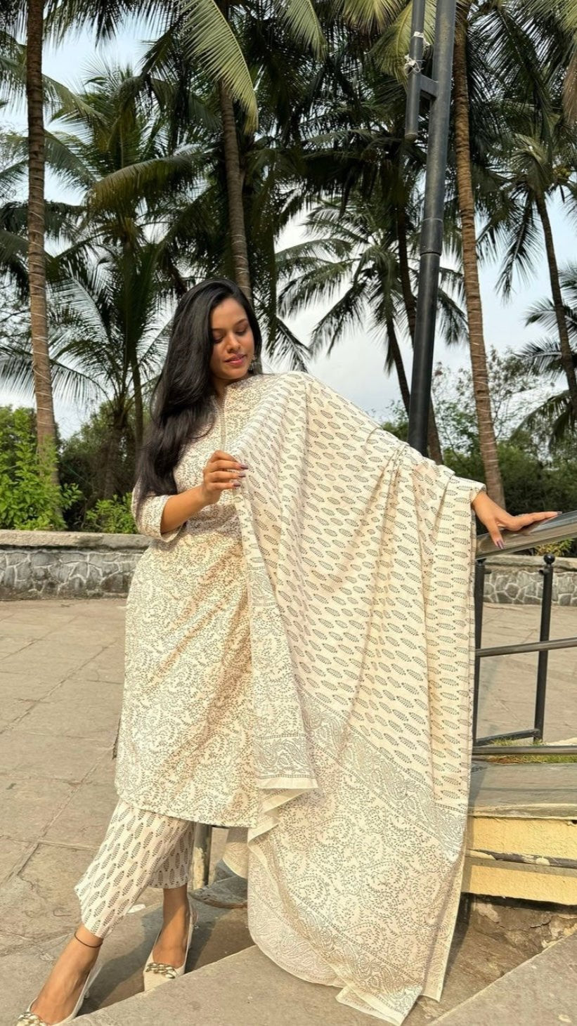 Woman wearing an elegant cream-white cotton kurta set with a light brown plant-inspired print, paired with a matching dupatta. She is posing outdoors against a background of lush green palm trees, exuding a blend of traditional ethnic style and natural charm.